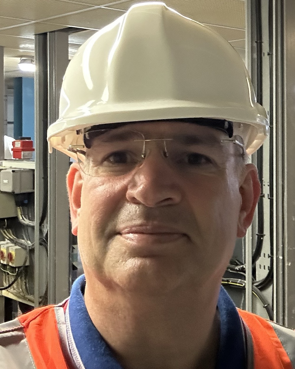 A man in his fifties in a white hard hat, safety glasses and an orange high-visibility vest over a navy shirt, with out-of-focus industrial control panels behind him.